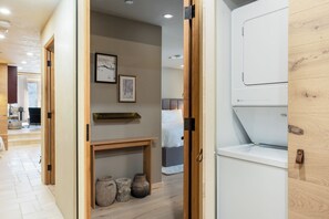 Une chambre d’hôtel avec une porte en bois, un lit, une table de chevet, une étagère avec des objets en céramique et un appareil électroménager blanc.