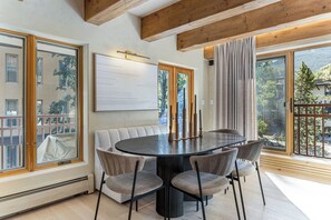 A dining area with a round table, cushioned chairs, and a view of the outdoors through large windows. - Village Center 3G | Renovated 2BR in the Heart of Vail Village (Vail)