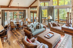 A spacious living room with a wooden floor, leather furniture, and large windows offering views of trees. - Gore Place | 5BR Luxury Vail Home, Hot Tub & Ski Slope Views (Vail)