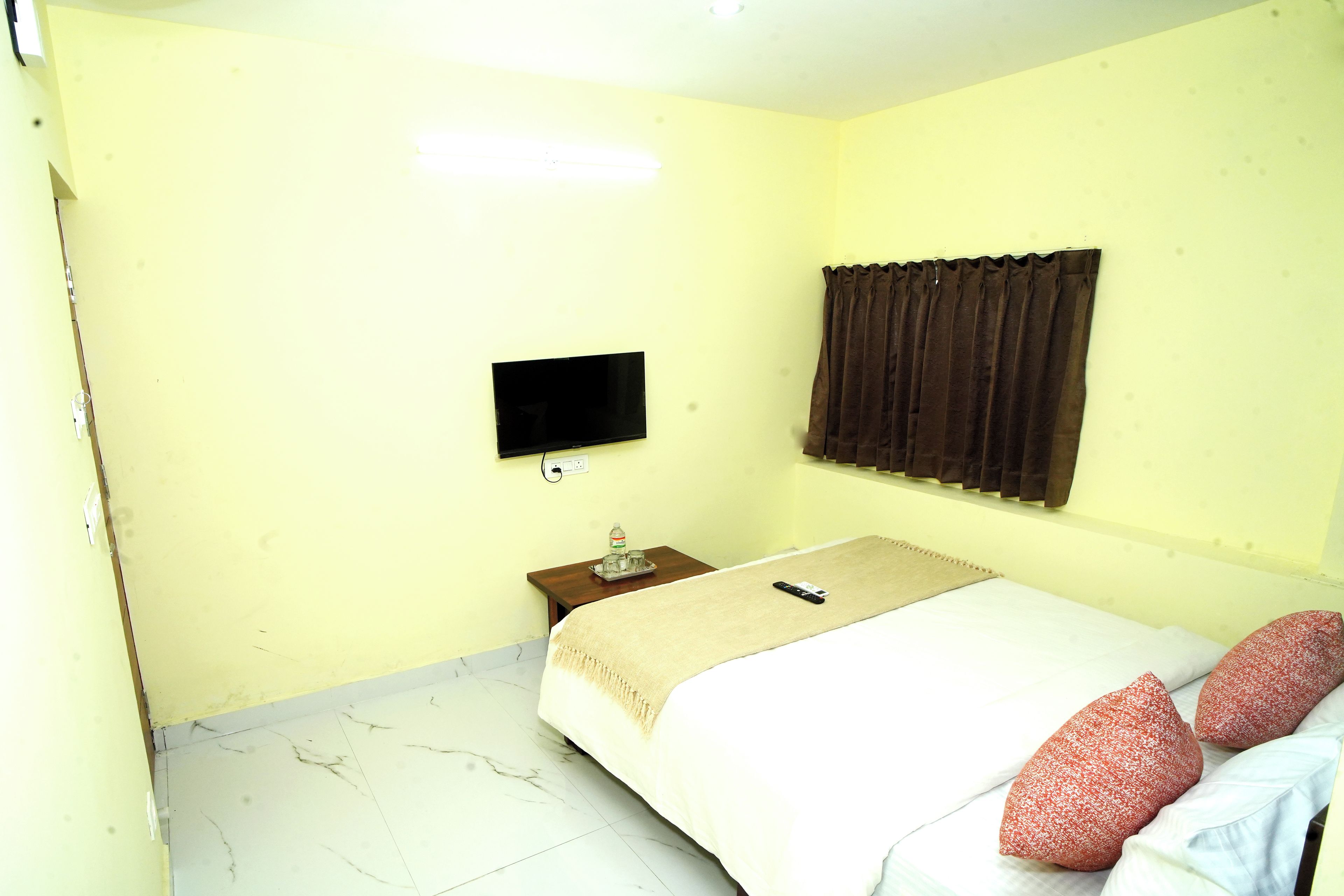 A small, simple room with a bed, a flat-screen TV mounted on the wall, a nightstand with a water bottle, and a curtain.