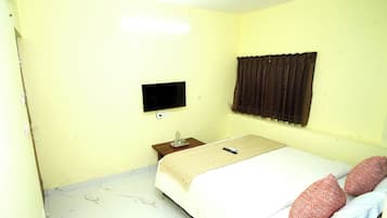 A small, simple room with a bed, a flat-screen TV mounted on the wall, a nightstand with a water bottle, and a curtain.