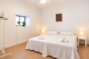 Une chambre d’hôtel avec un lit, deux tables de chevet, un ventilateur de plafond, une fenêtre avec des plantes et un panneau fixé au mur.