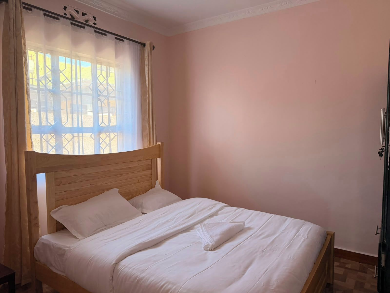 A single bed with a wooden headboard in a room with a plain wall and a window with a curtain.