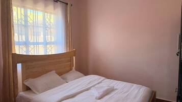 A single bed with a wooden headboard in a room with a plain wall and a window with a curtain.