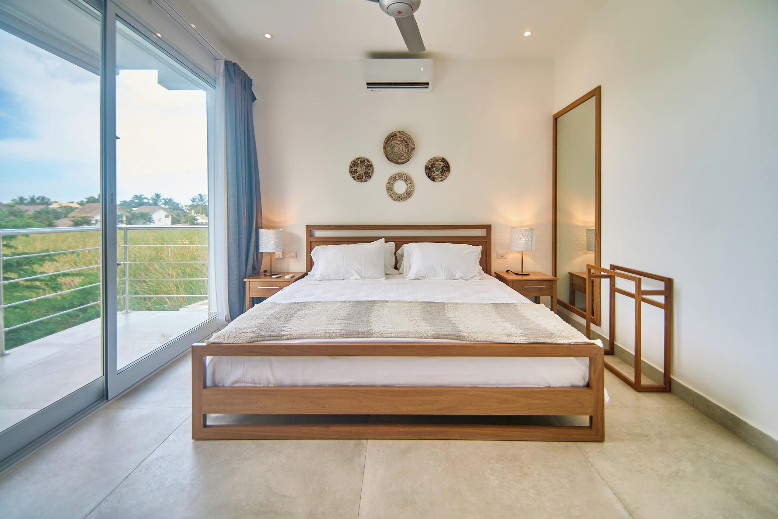 Quarto com cama grande, ventilador de teto, janela com vista para área verde e arte decorativa na parede.