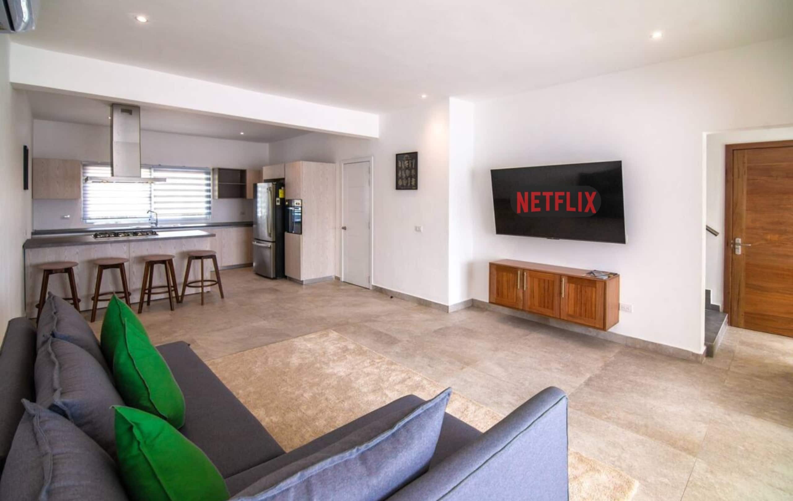 Een moderne woonkamer met een grijze bank, groene en blauwe kussens, een flatscreen televisie aan de muur en een keuken in de achtergrond.