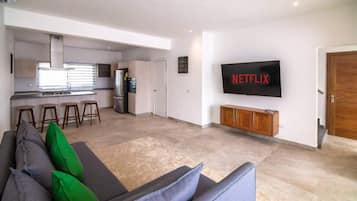 Een moderne woonkamer met een grijze bank, groene en blauwe kussens, een flatscreen televisie aan de muur en een keuken in de achtergrond.
