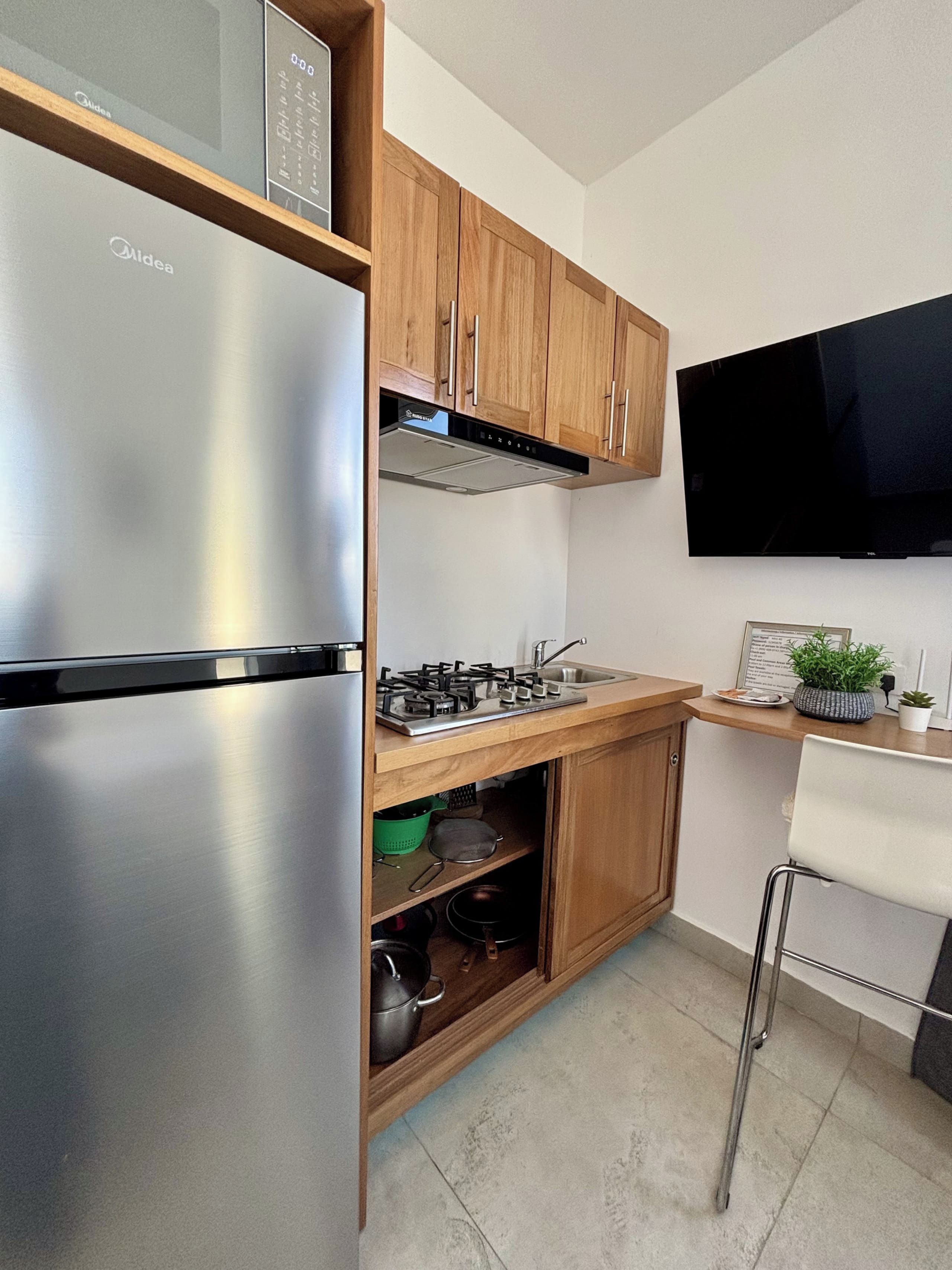 Cocina compacta con refrigerador de acero inoxidable, armarios de madera, estufa a gas y televisor de pantalla plana montado en la pared.