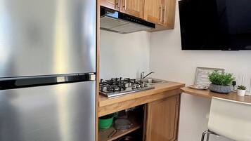 Cocina compacta con refrigerador de acero inoxidable, armarios de madera, estufa a gas y televisor de pantalla plana montado en la pared.