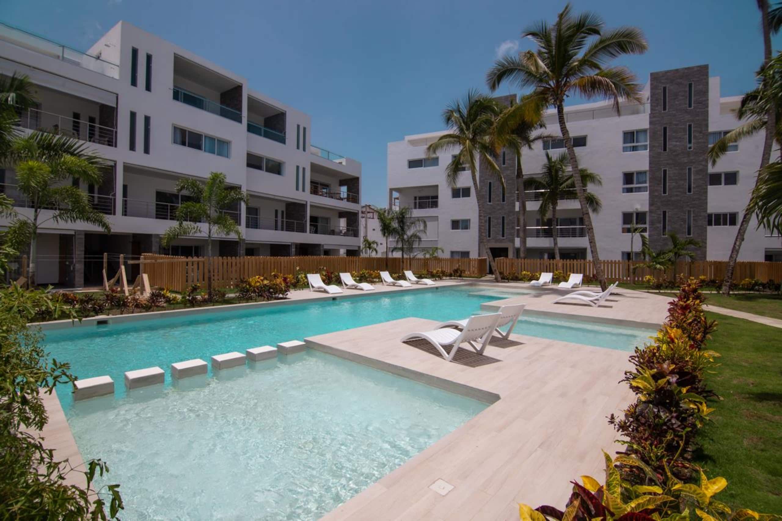 Un espace piscine moderne doté de nombreuses chaises longues et d’une piscine à l’eau d’un bleu limpide, le tout entouré d’immeubles à plusieurs étages dotés de balcons.