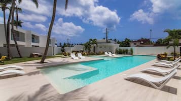 Una piscina rectangular con agua azul cristalina, rodeada de sillas de descanso blancas y palmeras.