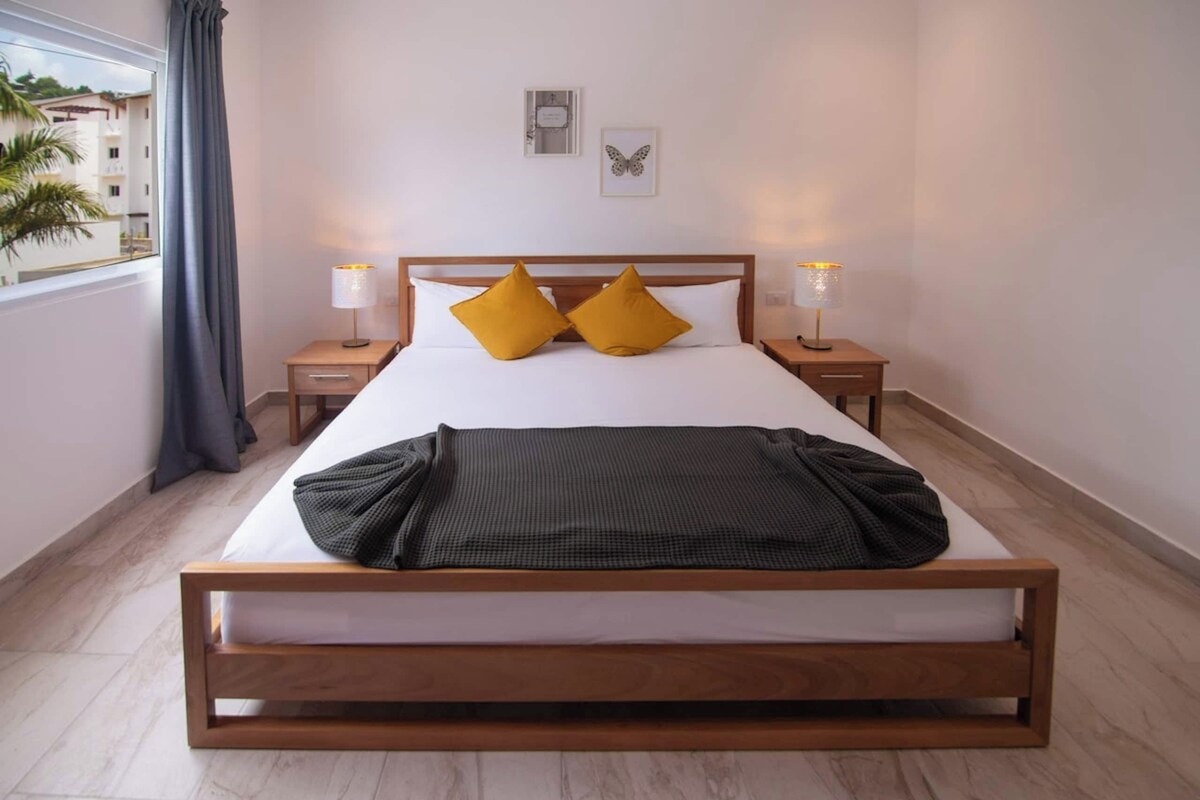 Habitación de hotel con una cama grande, dos lámparas de noche, una ventana con cortinas y vistas a edificios.