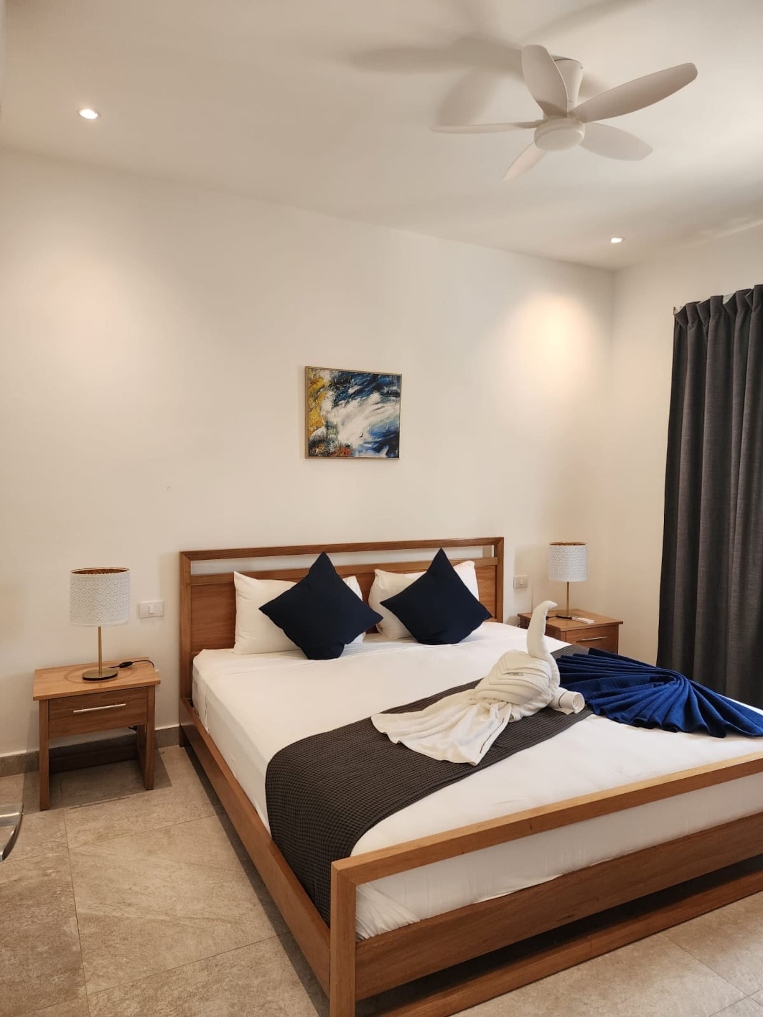 Habitación de hotel con cama, dos mesitas de noche, ventilador de techo, un cuadro y ventana con cortinas.