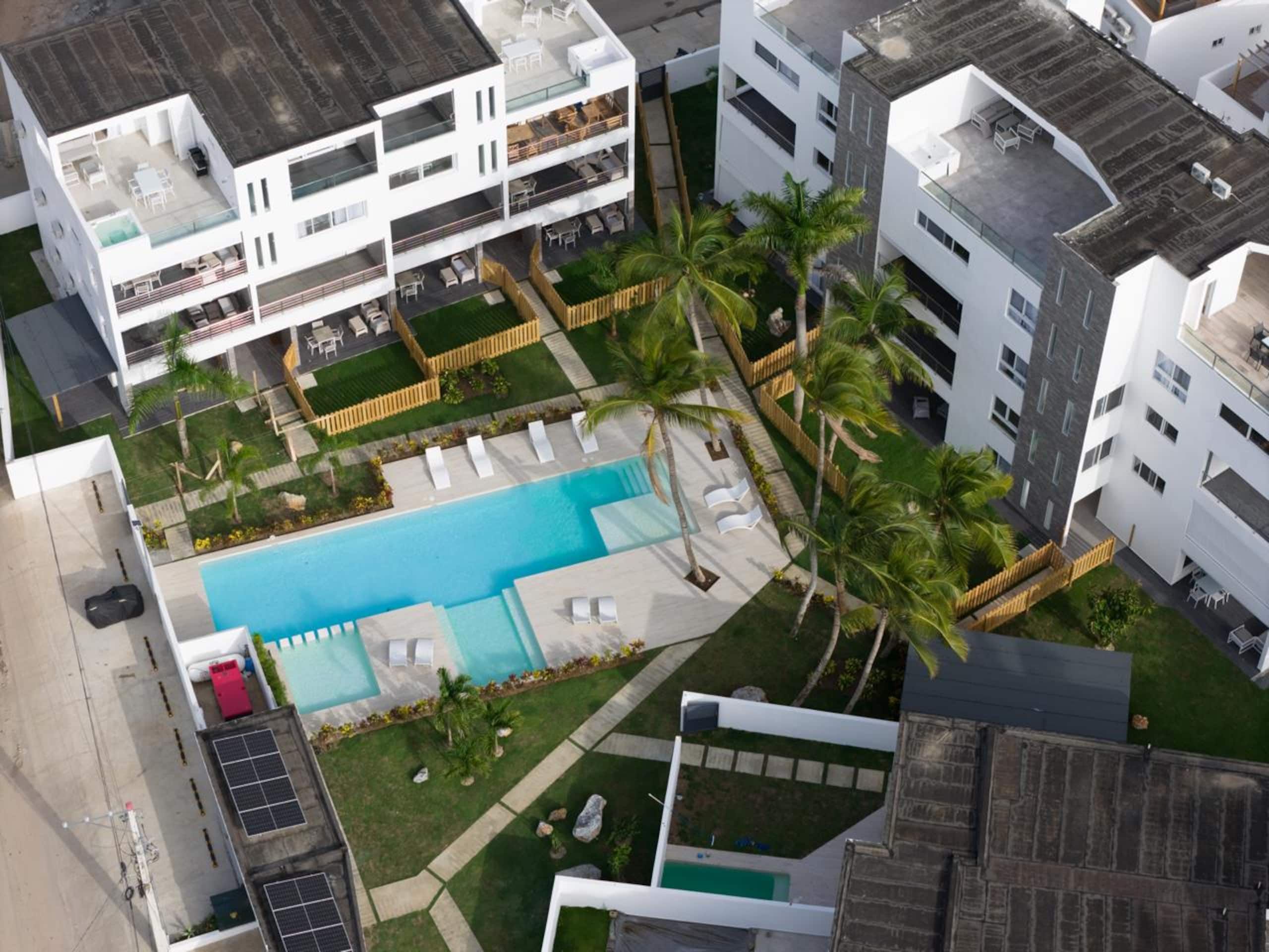 A modern residential building with a rooftop pool, surrounded by palm trees and a fenced garden area.
