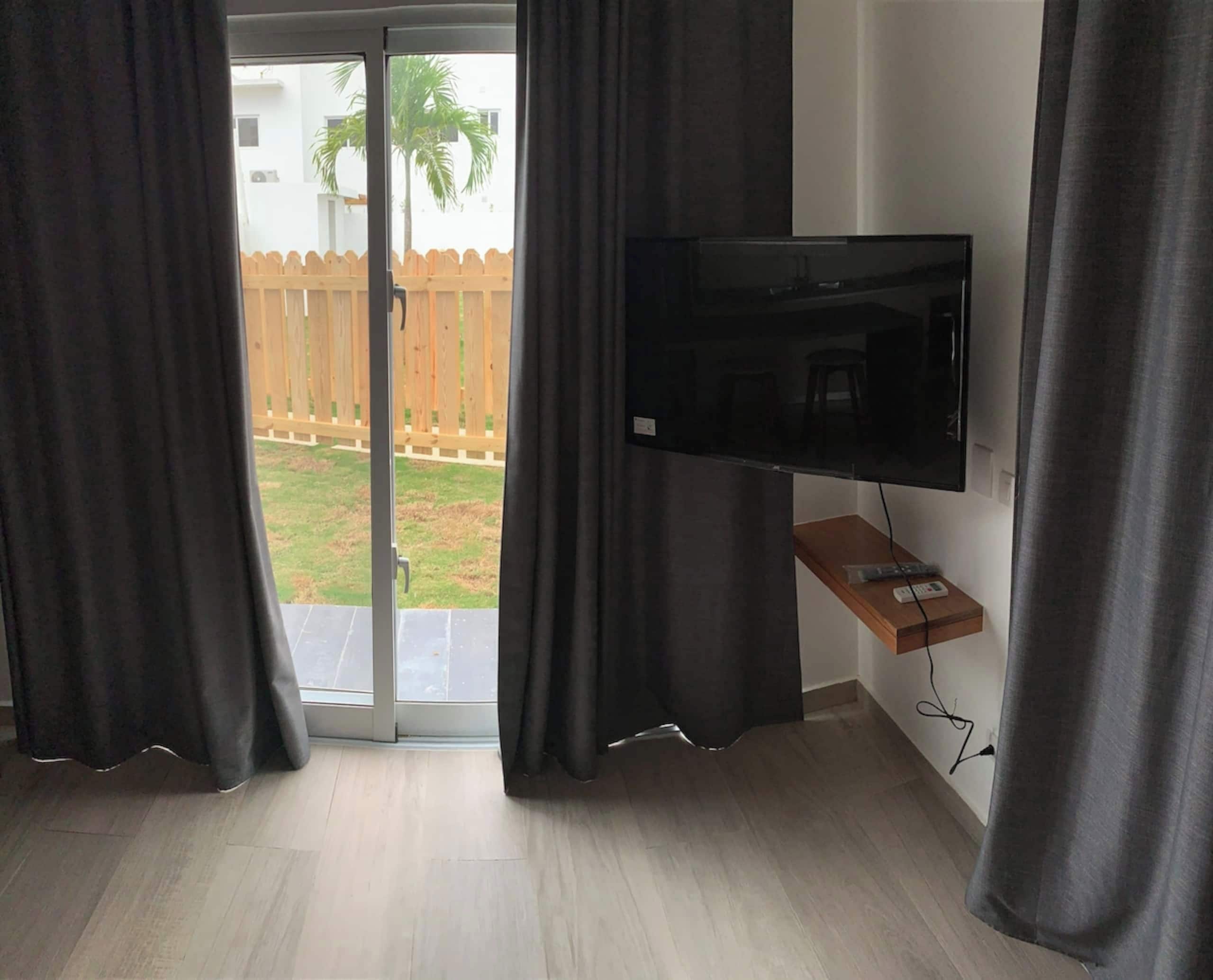 A room with a flat-screen TV mounted on a wall, a sliding glass door leading to a patio with a wooden fence and a palm tree, and a wooden floor.