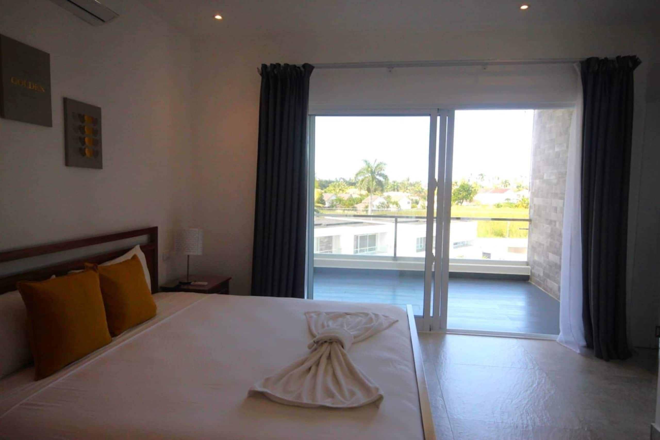 A hotel room with a large bed, a nightstand, a lamp, and a view of a pool and buildings through a sliding glass door.
