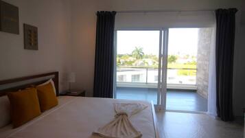 A hotel room with a large bed, a nightstand, a lamp, and a view of a pool and buildings through a sliding glass door.