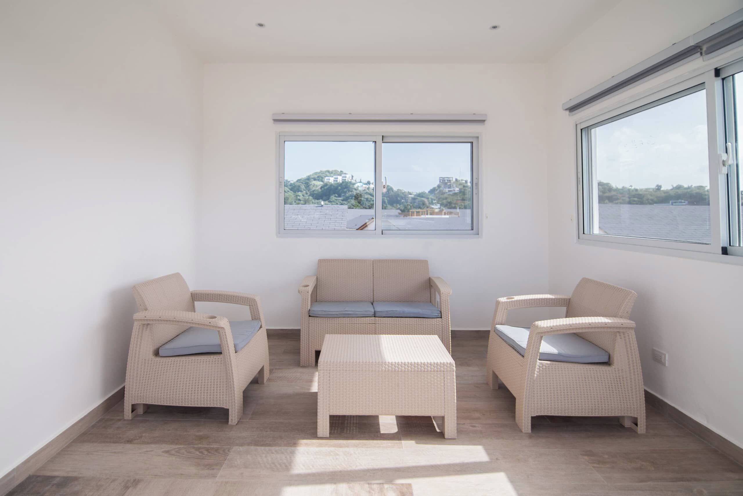 Ein Zimmer mit einer Couch, zwei Sesseln und einem kleinen Tisch. Zwei Fenster bieten Blick auf ein Gewässer und Hügel.