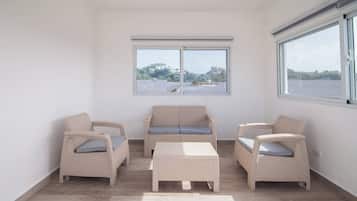 Ein Zimmer mit einer Couch, zwei Sesseln und einem kleinen Tisch. Zwei Fenster bieten Blick auf ein Gewässer und Hügel.