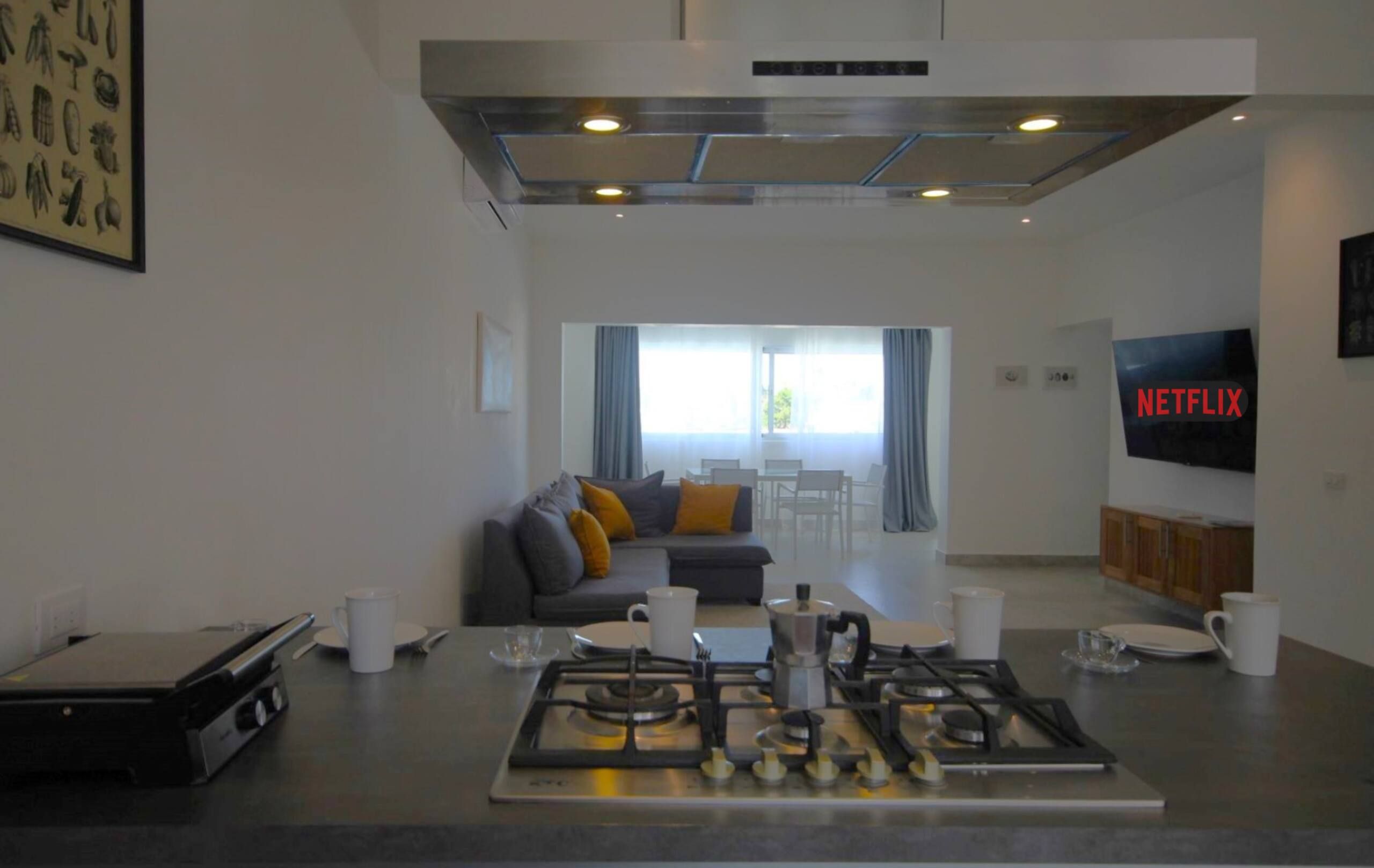 A modern kitchen with a gas stove, a coffee machine, and a dining area with a sofa and a TV displaying the Netflix logo.