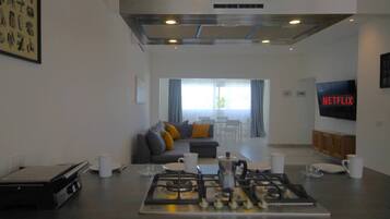 A modern kitchen with a gas stove, a coffee machine, and a dining area with a sofa and a TV displaying the Netflix logo.