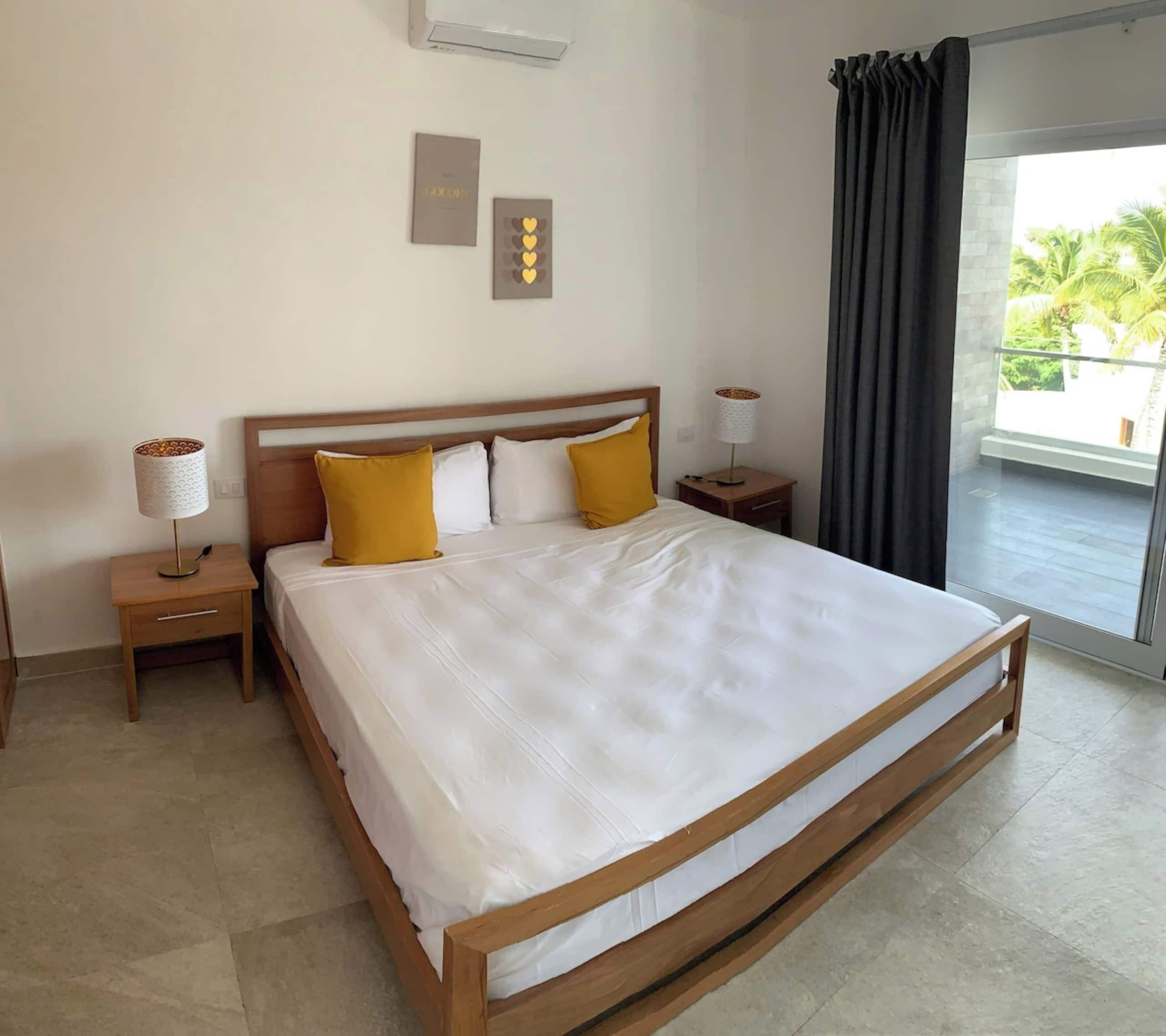A hotel room with a large bed, two bedside tables, a lamp, a wall-mounted air conditioner, and a view of the outdoors.