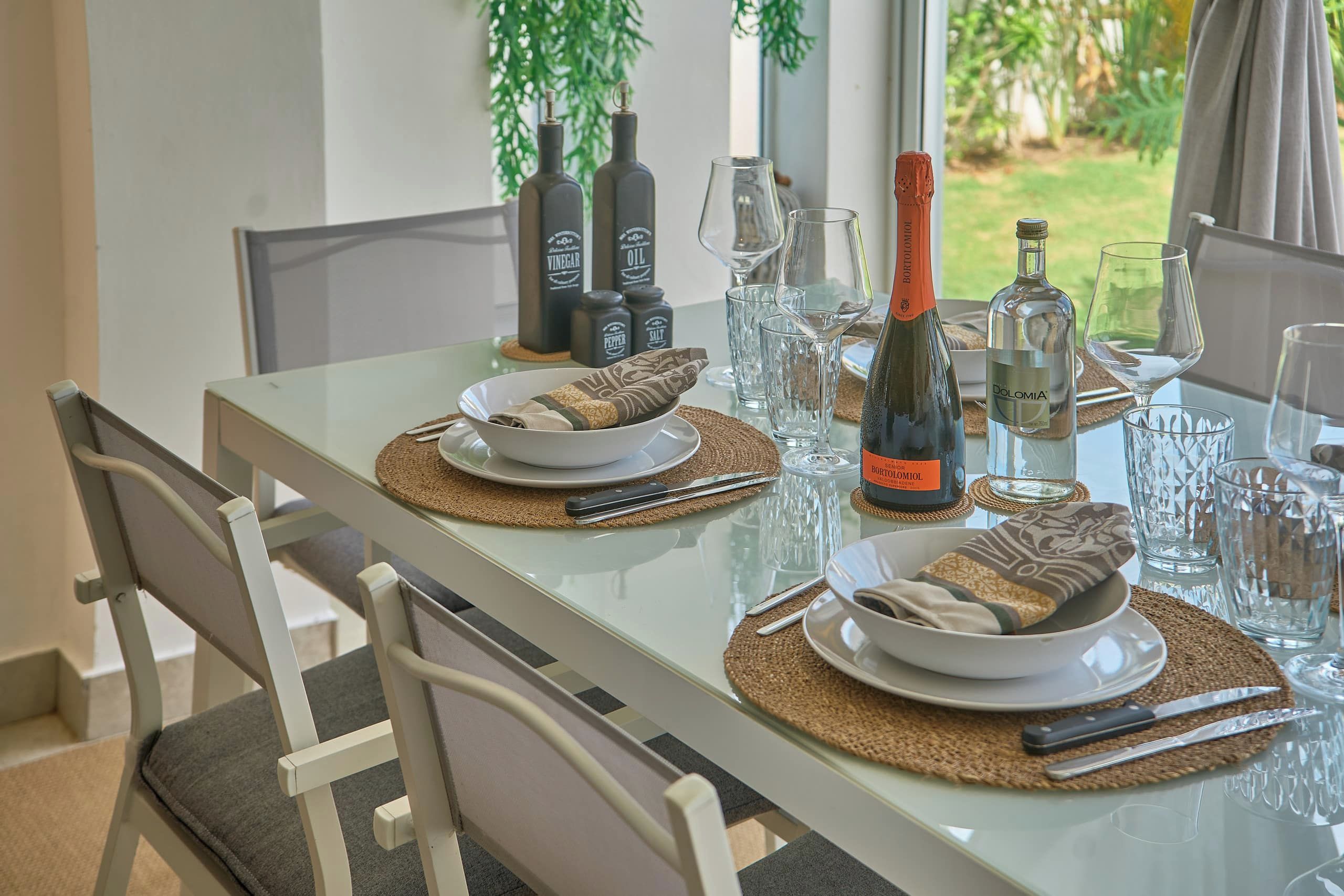 Mesa de comedor dispuesta con platos, cubiertos y copas, con botellas de bebidas y salsas de mesa al fondo.