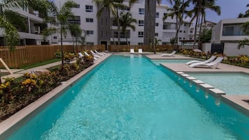 Una piscina rectangular con agua cristalina azul, rodeada de sillas de descanso blancas y palmeras.