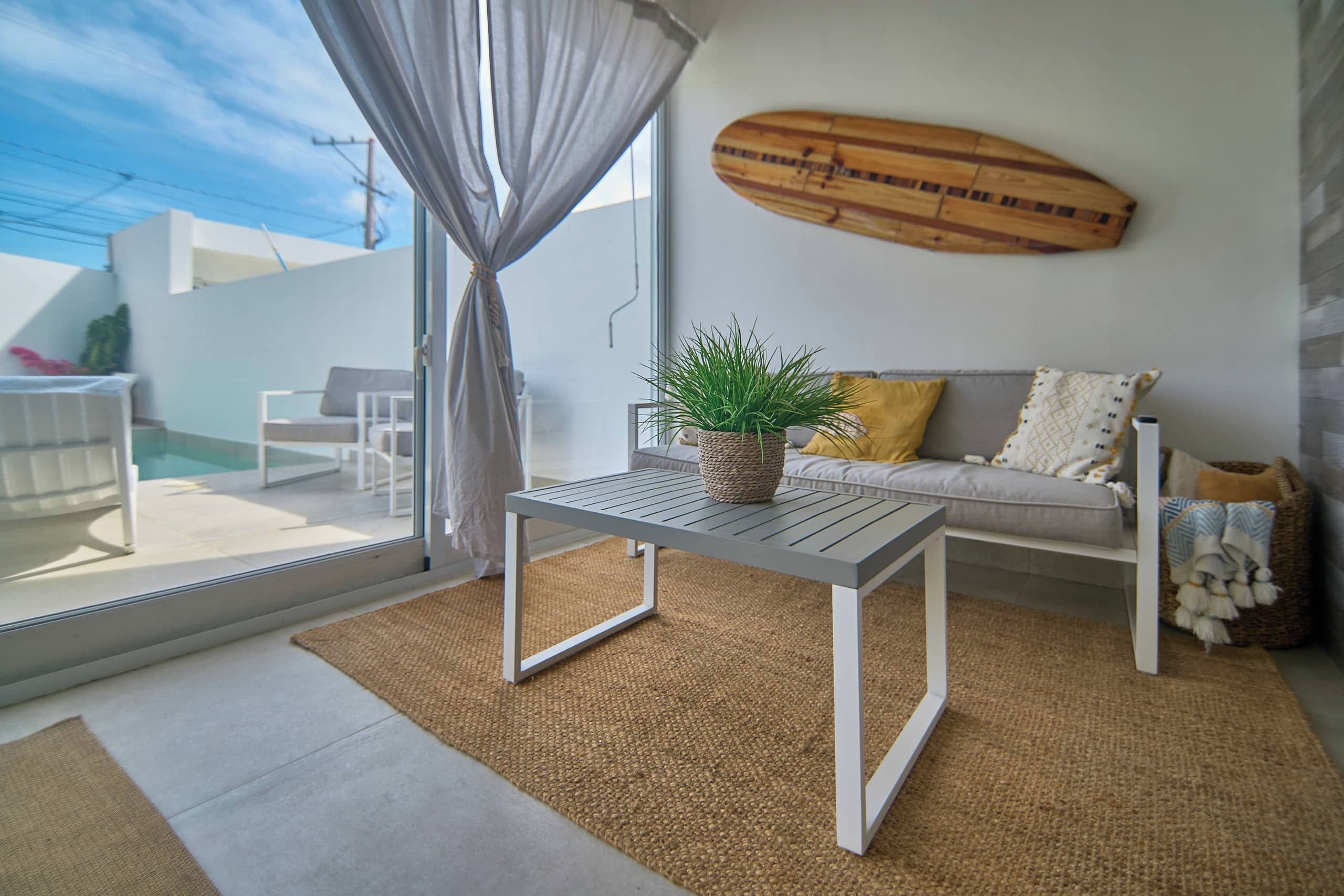 A modern living room with a sofa, a table with a plant, and a surfboard on the wall.