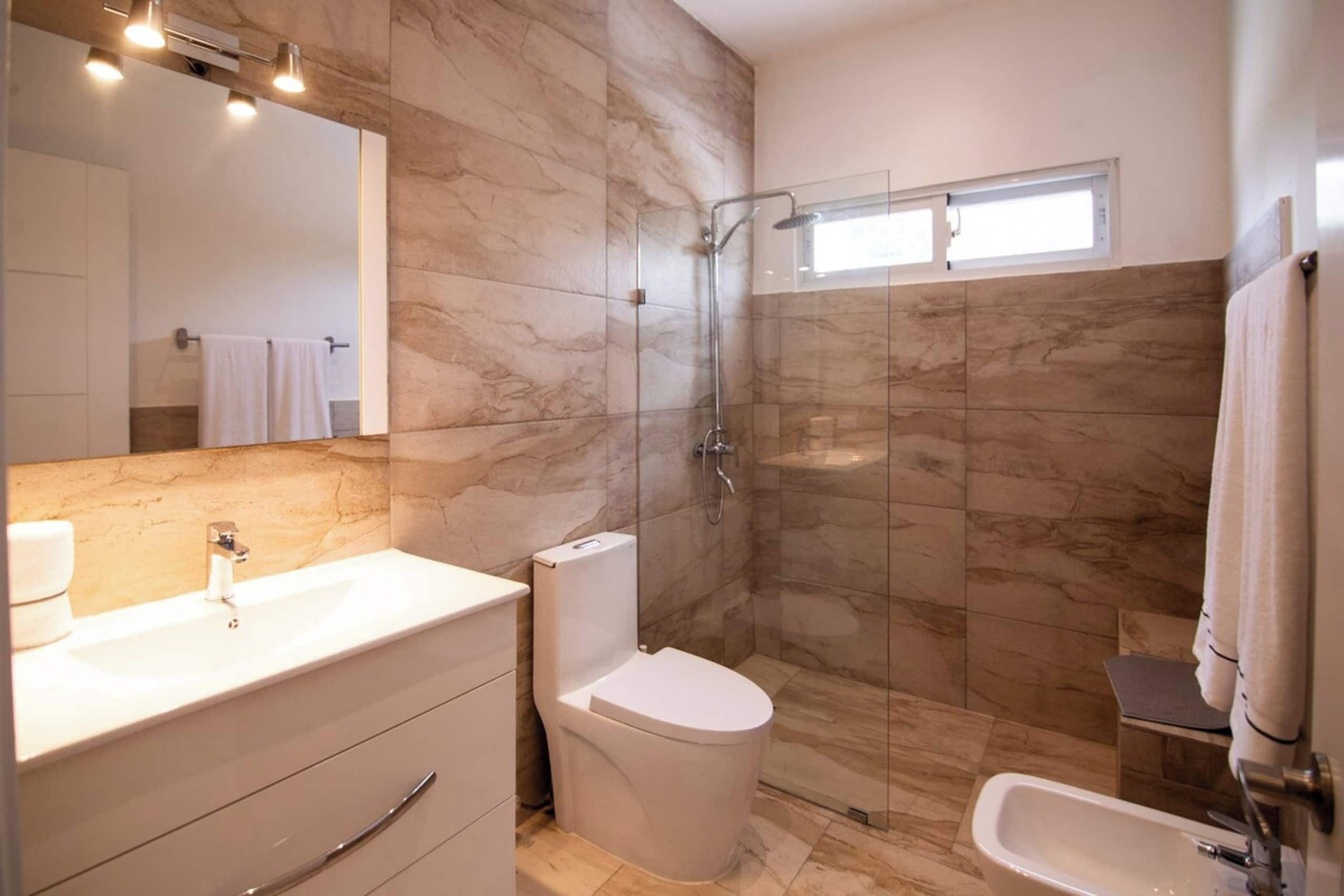 A modern bathroom with a marble wall, a glass shower enclosure, a white toilet, and a sink with a mirror above it.