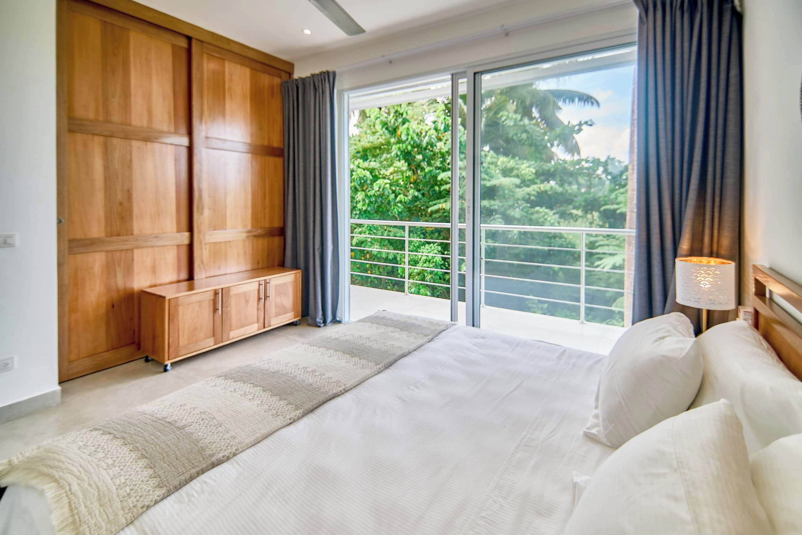 Une chambre avec un grand lit, une armoire en bois, un balcon donnant sur un paysage verdoyant et une table de chevet avec une lampe.