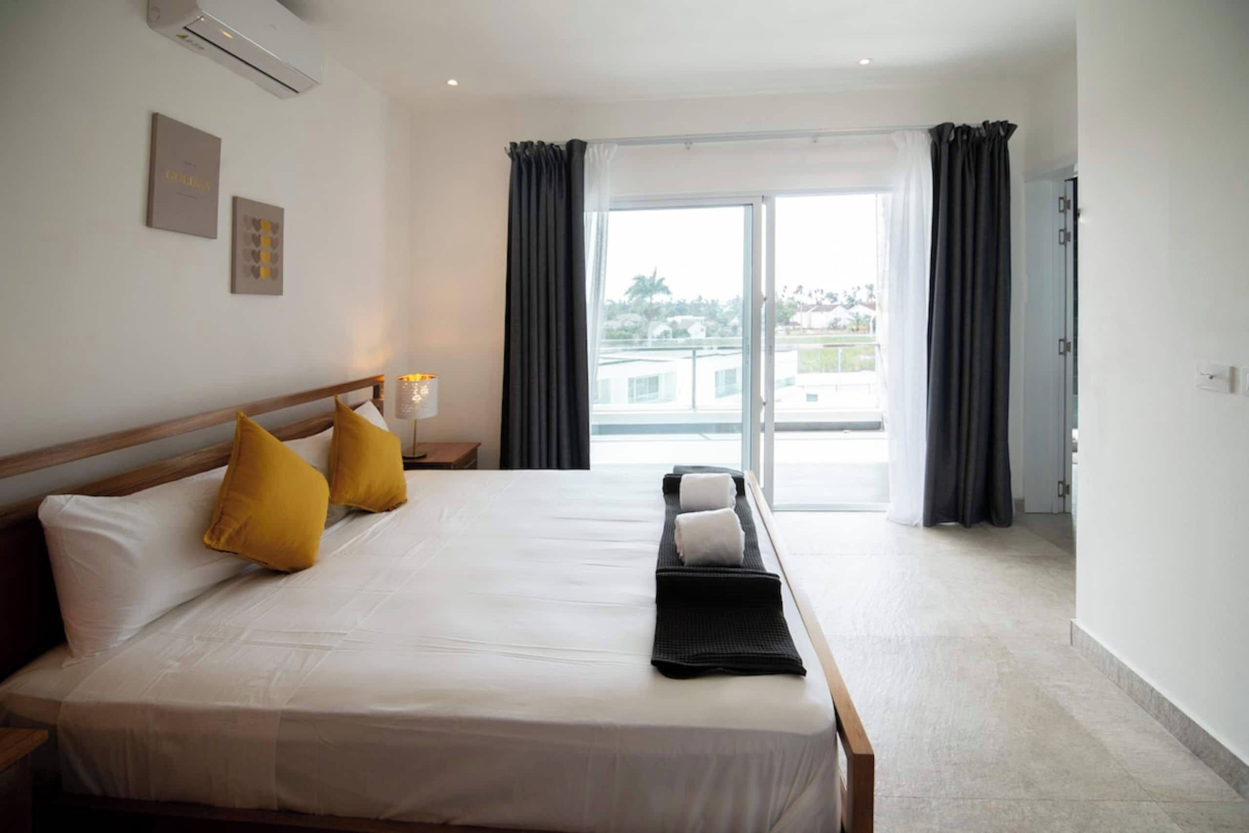 Ein Hotelzimmer mit Bett, Kissen, Nachttisch, Lampe und Blick ins Freie.