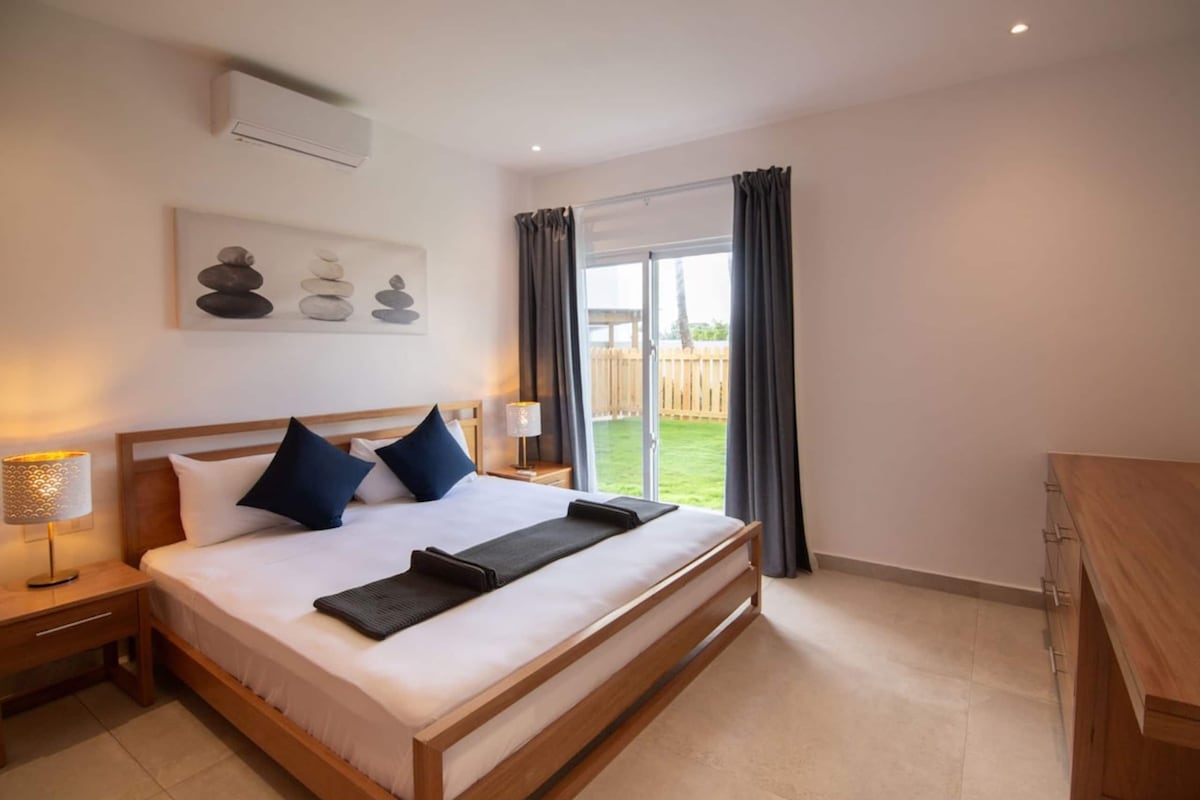 Habitación de hotel con una cama grande, una mesita de noche con lámpara, vista al exterior a través de una puerta corrediza y un equipo de aire acondicionado instalado en la pared.