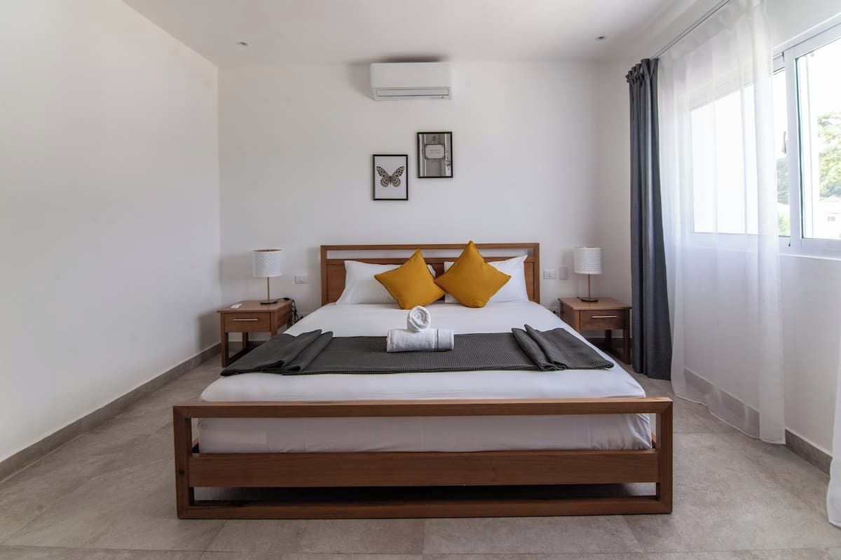 A bedroom with a wooden bed, two bedside tables, a wall-mounted air conditioner, and a window with curtains.