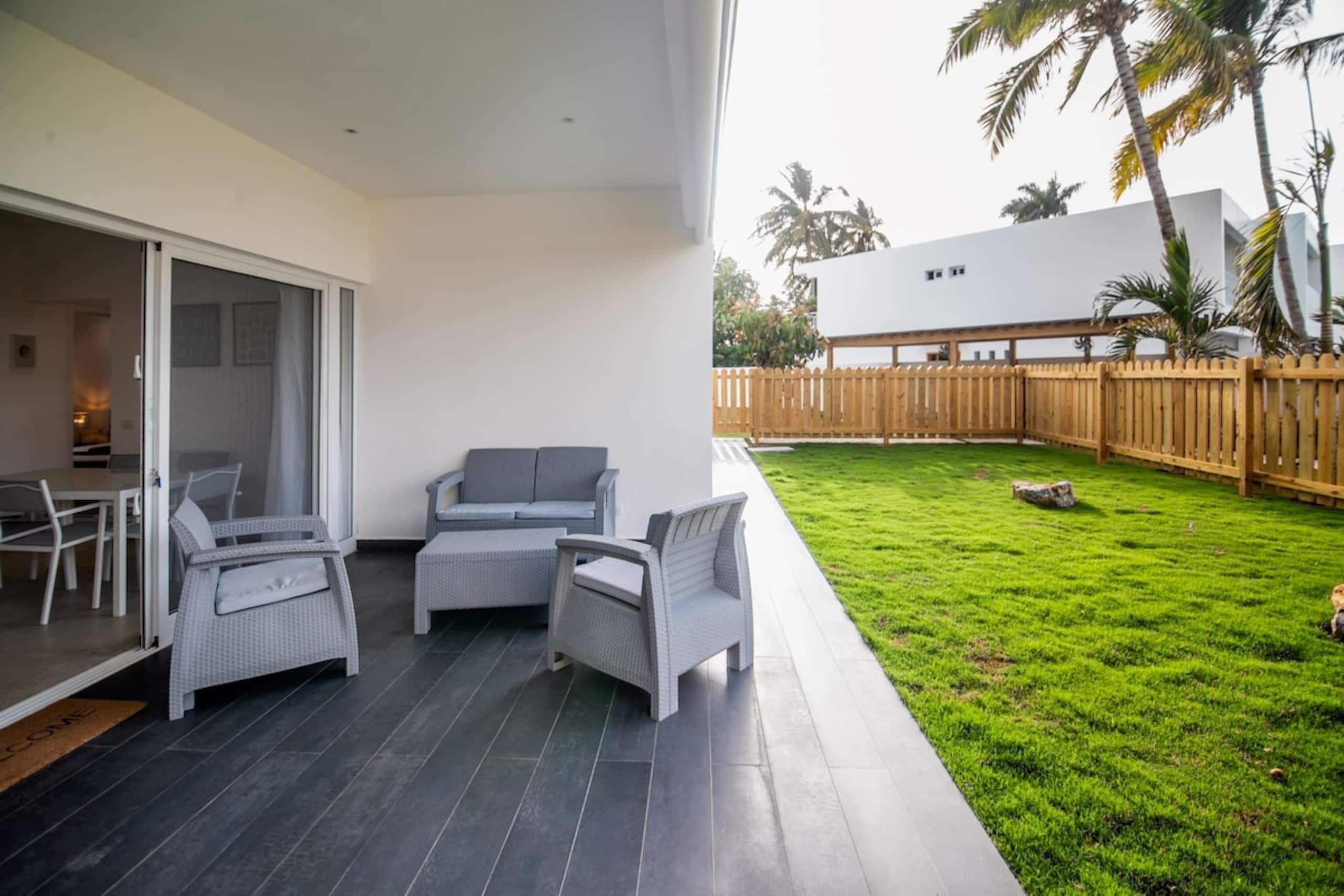 Un patio moderno al aire libre con mobiliario de ratán gris, puerta de vidrio y césped cuidado con valla de madera.
