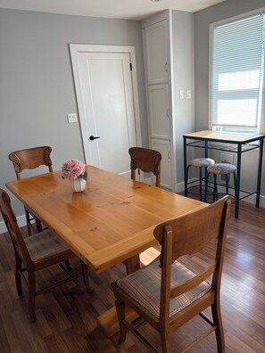 A dining area with a wooden table and four chairs, a vase with flowers, and a window with blinds.