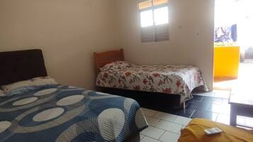 A room with two beds, one with a blue and white patterned bedspread and the other with a floral bedspread. There is a small window and a door leading outside.