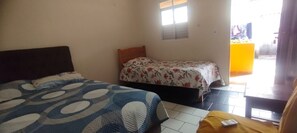 A room with two beds, one with a blue and white patterned bedspread and the other with a floral bedspread. There is a small window and a door leading outside.