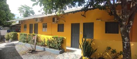 A yellow building with a tiled roof, a door, and windows. There are plants and a tree in the foreground.