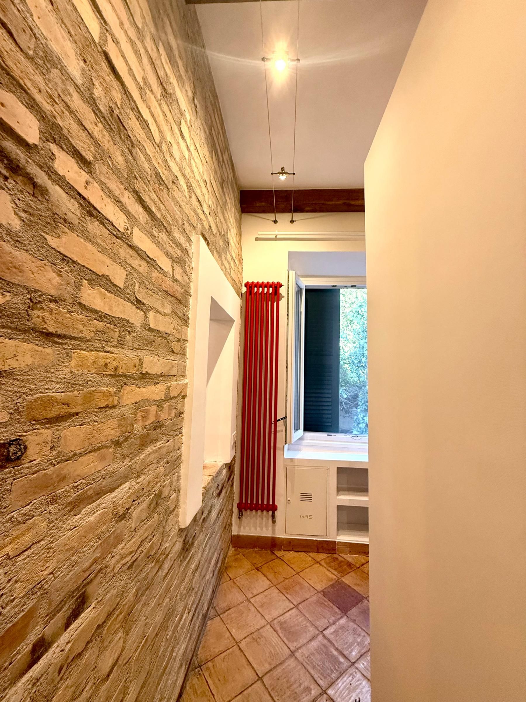 Un couloir étroit avec un mur en briques, une fenêtre ornée de rideaux rouges et un sol carrelé.