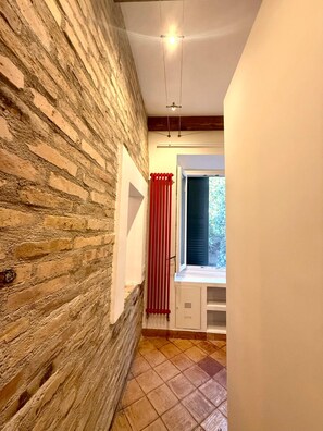 Un couloir étroit avec un mur en briques, une fenêtre ornée de rideaux rouges et un sol carrelé.