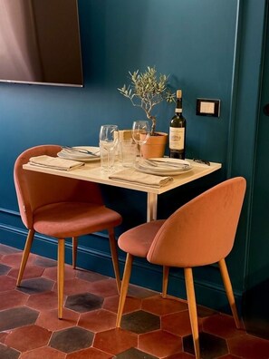 Een eettafel gedekt met borden, glazen en een fles, met aan weerszijden twee stoelen, tegen een blauwe muur met een potplant.