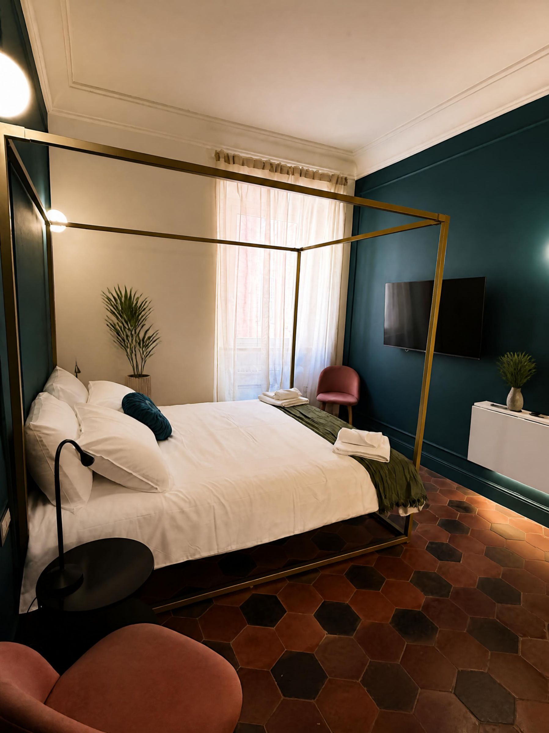 A modern bedroom with a four-poster bed, a flat-screen TV mounted on the wall, and a hexagonal tiled floor.