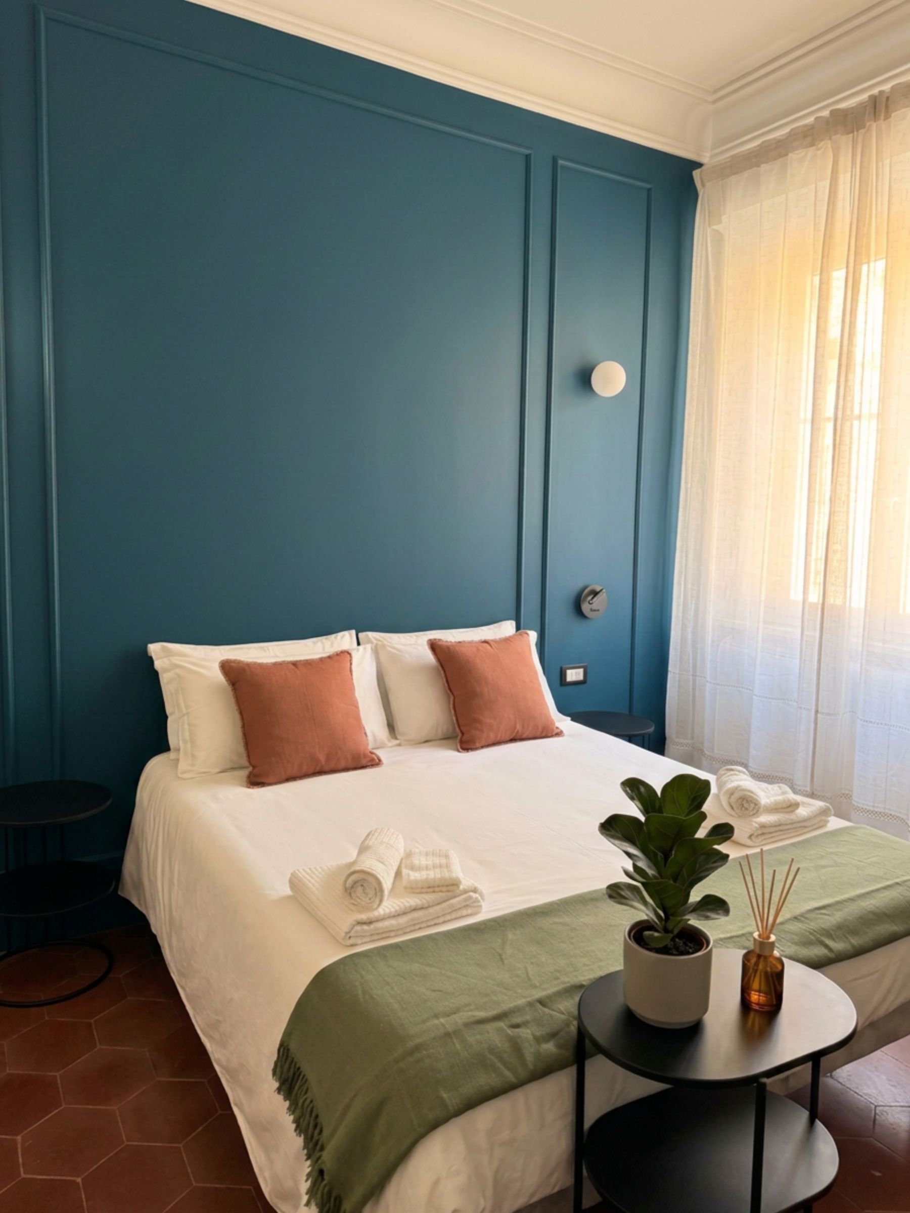Una camera da letto con un letto, cuscini, un comodino con una pianta e una finestra con tende.