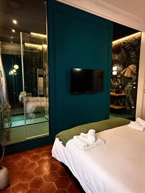 Una camera d'albergo moderna con un letto grande, una televisione a schermo piatto montata su una parete color verde acqua e un box doccia in vetro, arricchita da un motivo decorativo a tema tropicale.