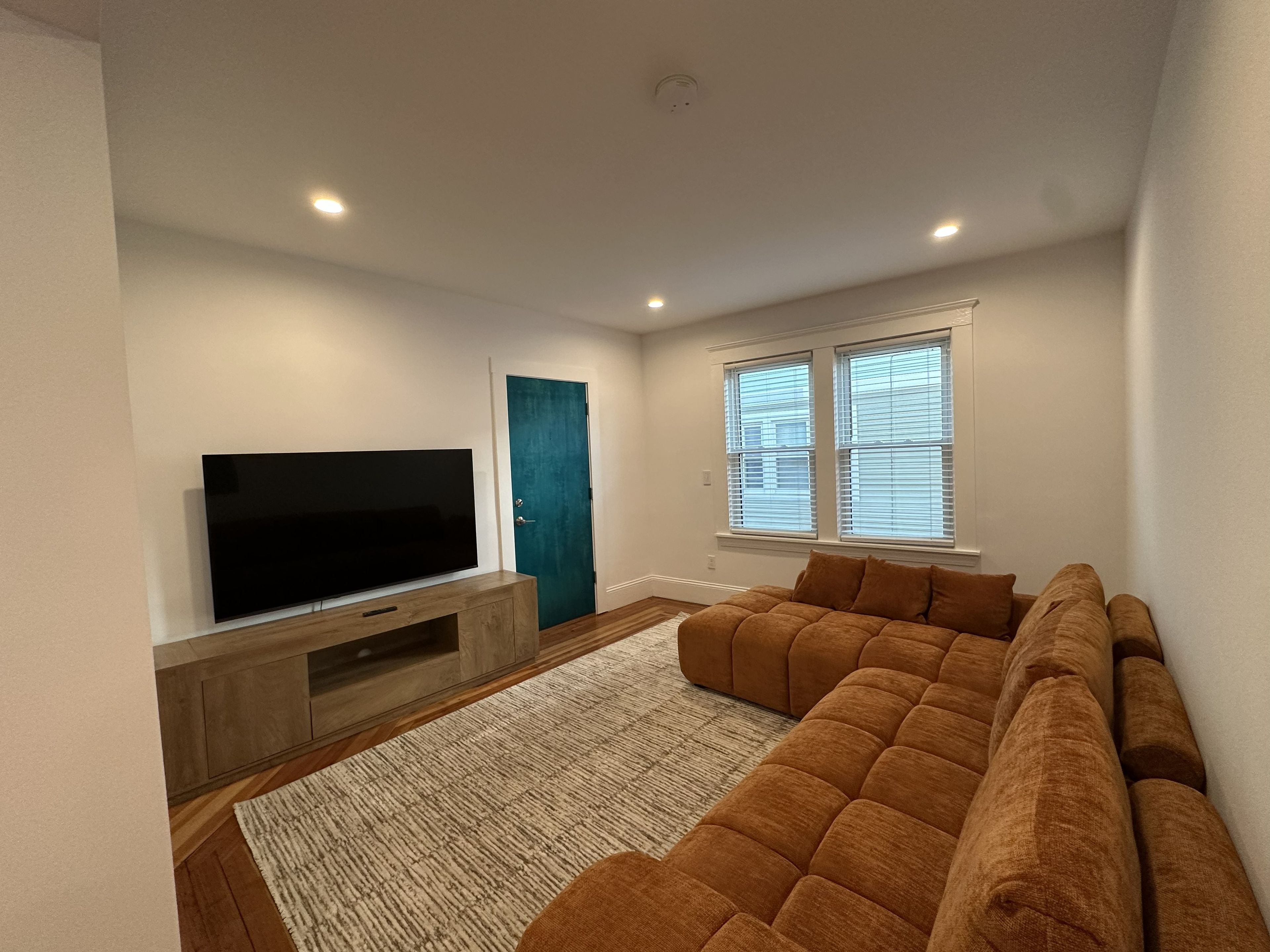 Een woonkamer met een flatscreen televisie op een houten standaard, een blauwgroene deur en een grote bruine bank.