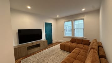 Een woonkamer met een flatscreen televisie op een houten standaard, een blauwgroene deur en een grote bruine bank.
