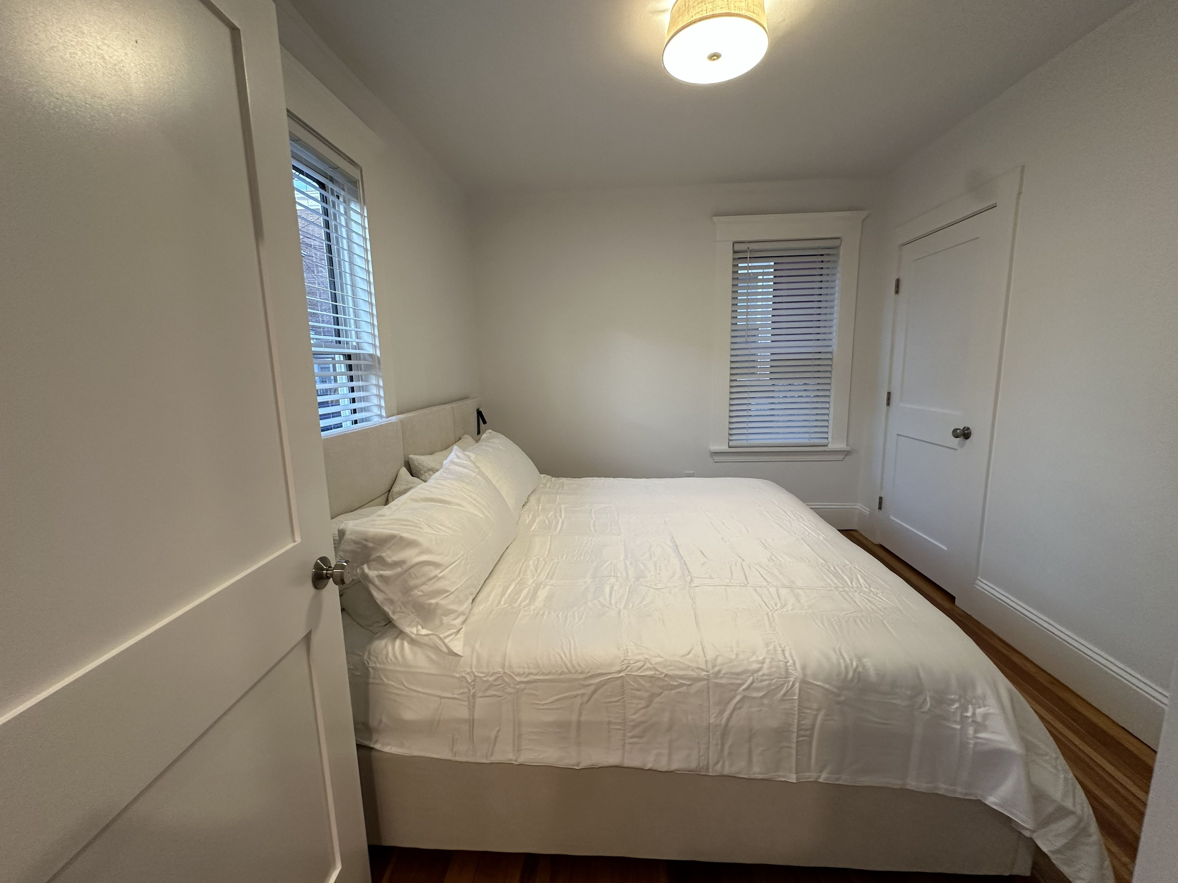 A bedroom with a bed, two windows with blinds, a door, and a ceiling light fixture.