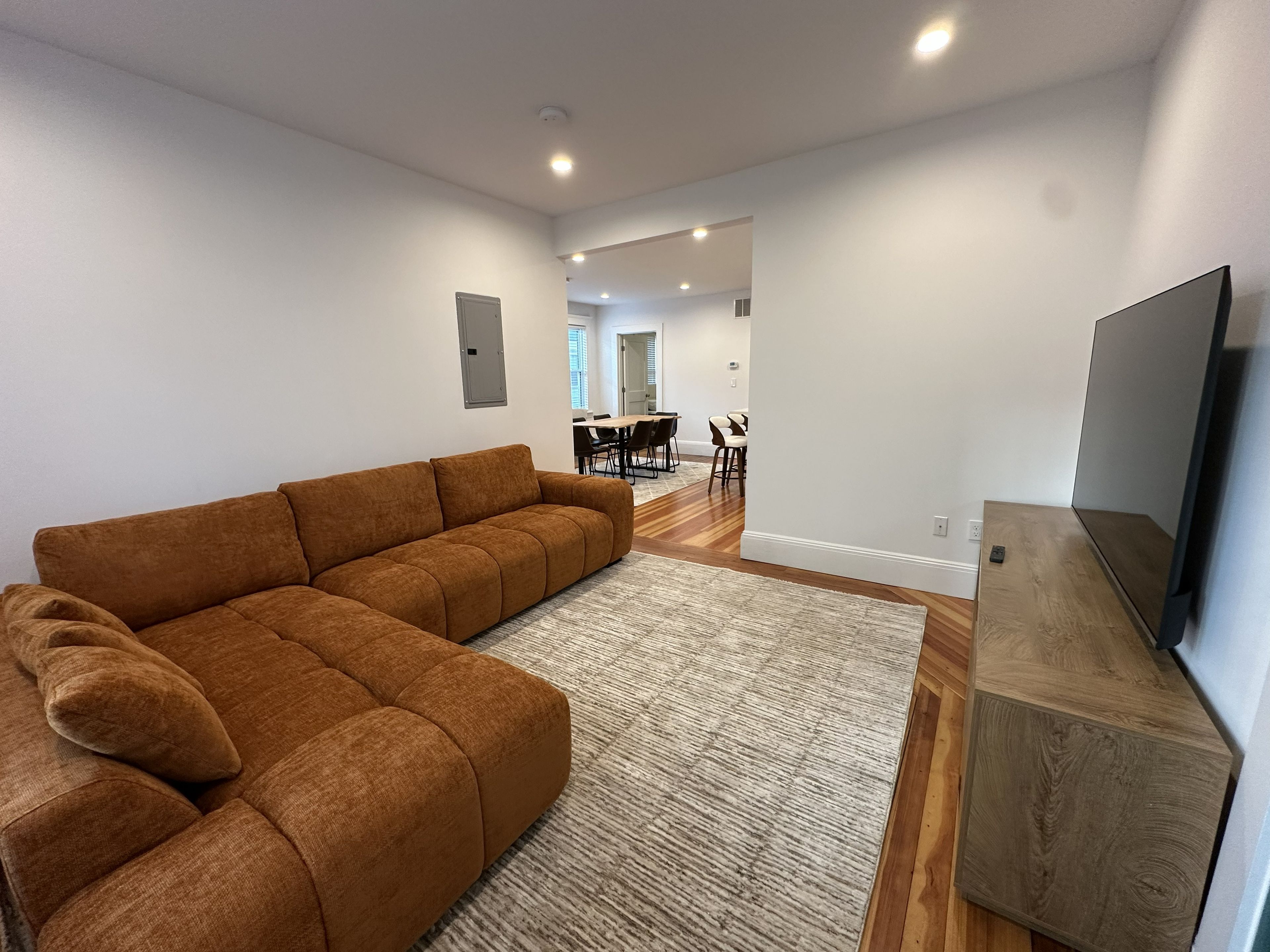 A modern living room with a sectional sofa, a flat-screen TV, and a dining area in the background.
