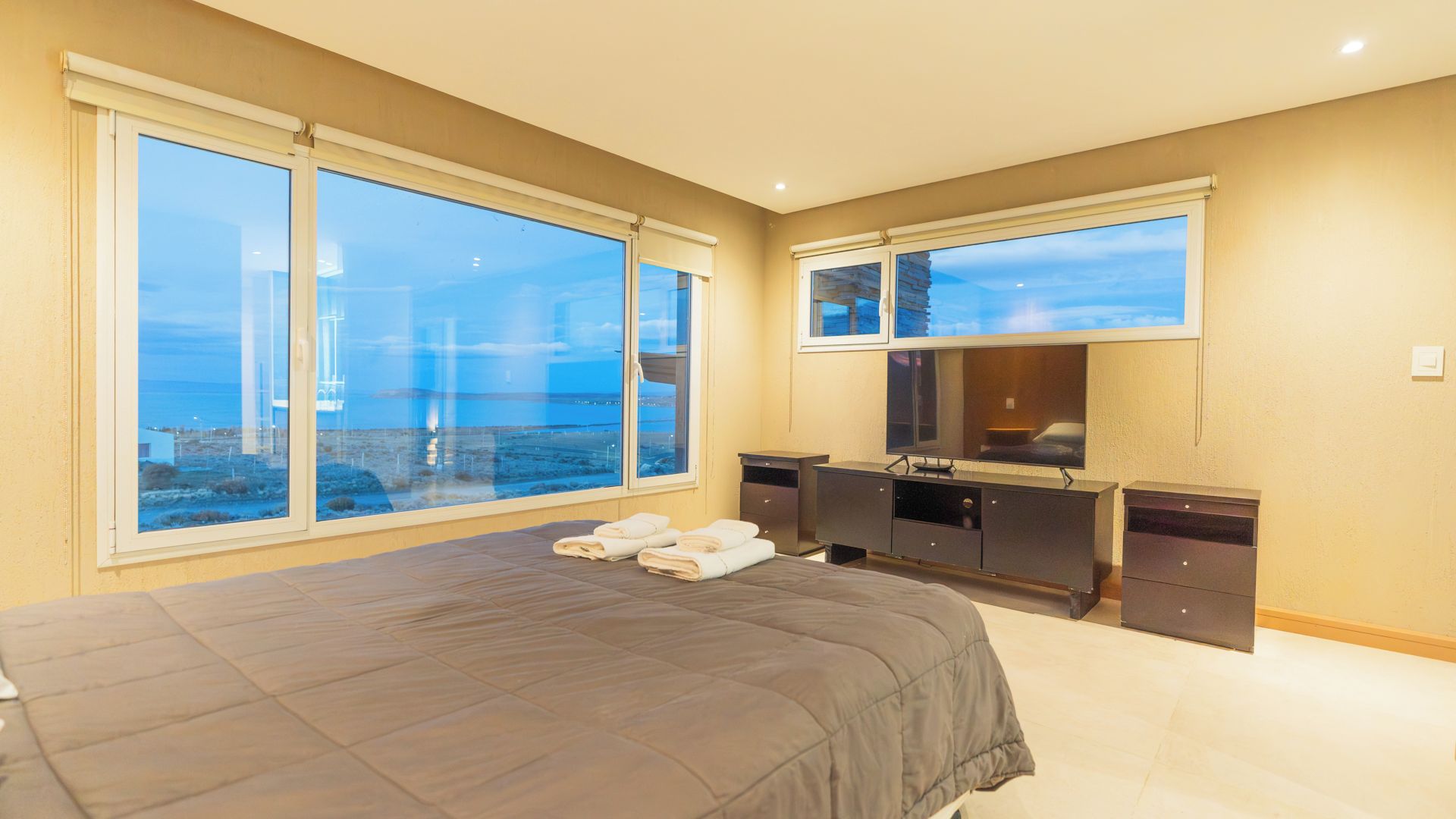 A bedroom with a large bed, a dresser, a mirror, and a view of the cityscape through large windows.